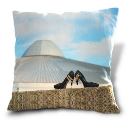 An image of a cushion, the cushion having a cover showing a pair of black high heel shoes sat on a wall in front of a building with blue sky behind it