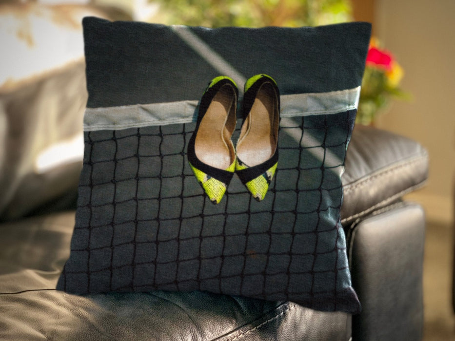 An image of a cushion on a couch, the cushion having a cover showing a pair of yellow high heel shoes hung on a tennis net