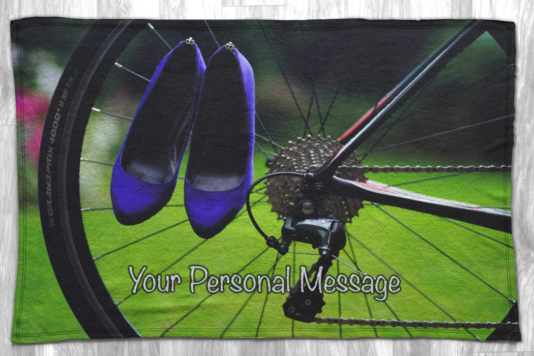 a fleece blanket flat on a wooden floor, the blanket having an image of a rear of a bicycle with a pair of ladies shoes hung over the wheel, with a personal message printed