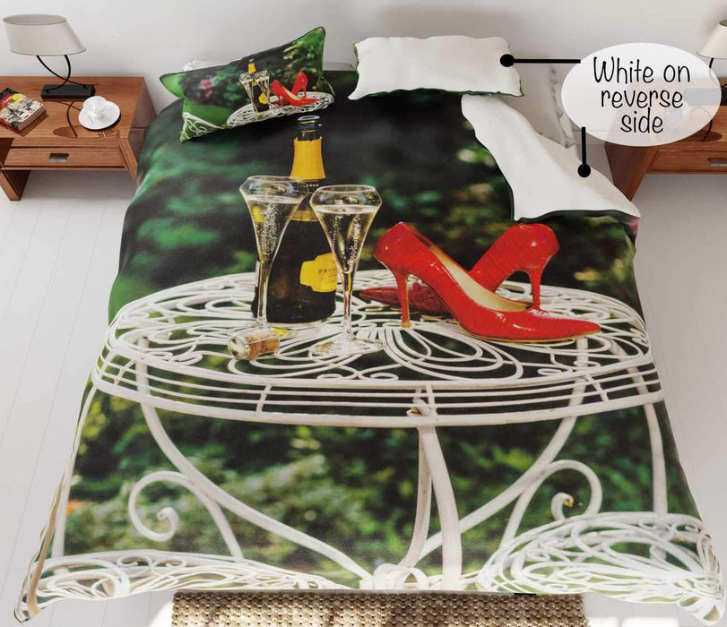 A duvet cover on the bed,the duvet having an image of a bottle of fizzy wine with two poured glasses upon a garden table, along with a pair of red high heel shoes on the table, the duvet is folded to show a white underside