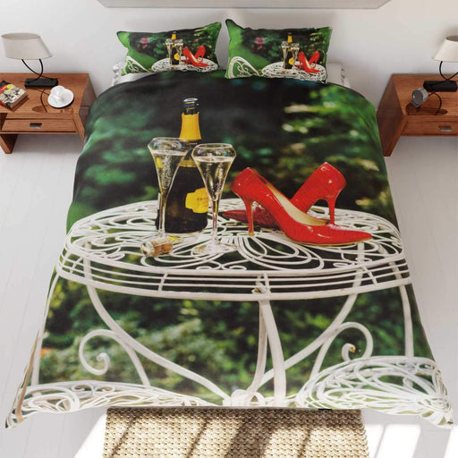 A duvet cover on the bed,the duvet having an image of a bottle of fizzy wine with two poured glasses upon a garden table, along with a pair of red high heel shoes on the same table
