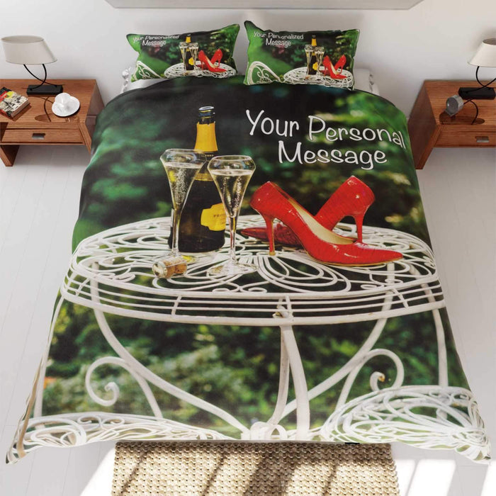 A duvet cover on the bed,the duvet having an image of a bottle of fizzy wine with two poured glasses upon a garden table, along with a pair of red high heel shoes on the same table, and a personal message