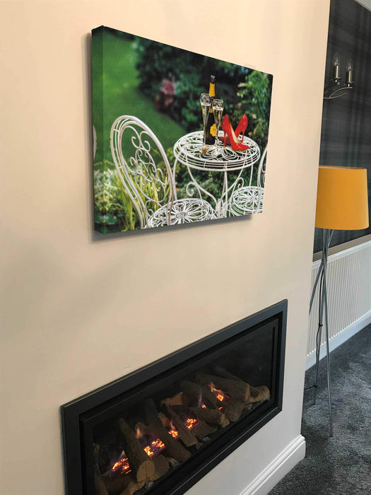 A canvas print hung above a fireplace, the canvas print having an image of a pair of red high heel shoes on a table in a garden, there is a bottle of fizzy wine and two filled glasses next to the shoes