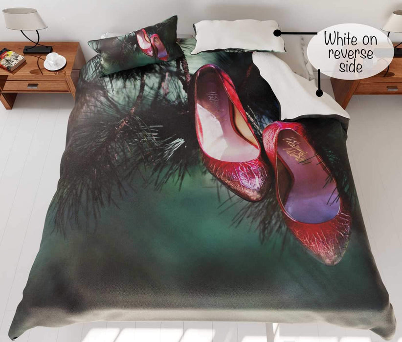 A duvet cover on a bed, the duvet cover having image of a red pair of high heel shoes hung from branch of a tree, a corner of the duvet is folded back to show a white underside