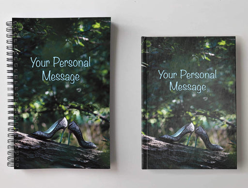 Two notebooks, side by side, both having covers with image of a tree within a forest, upon a think branch of the tree is a pair of ladies high heel shoes, with a personal message on the cover