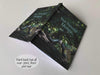An open hardback notebook having a cover with image of a tree within a forest, upon a think branch of the tree is a pair of ladies high heel shoes, with a personal message on the cover