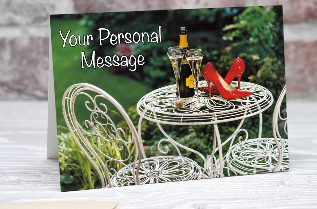 A birthday, greetings of invitation card showing a pair of red shoes on a garden table along side a glass and bottle of sparkling wine set within a garden setting, along with a personal message