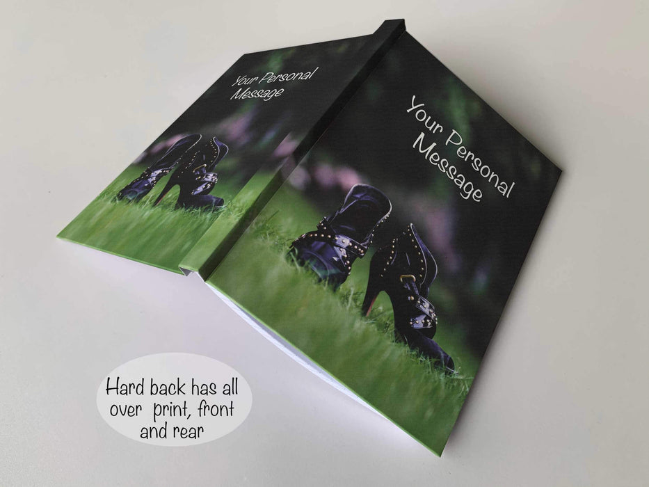 An open hardback notebook having a cover with an image of a close up image of a pair of purple ladies high heel ankle boots in middle of a garden, with a personal message on the cover