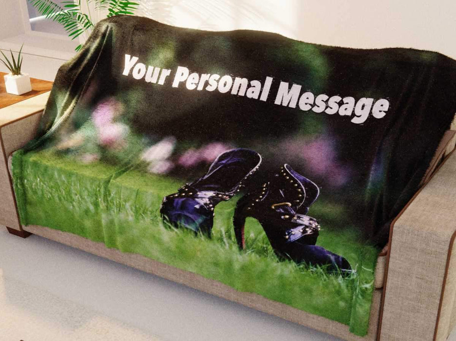 A blanket draped over a couch, the blanket having an image of a pair of purple high heel shoes sat on a grass lawn, along with a printed personal message