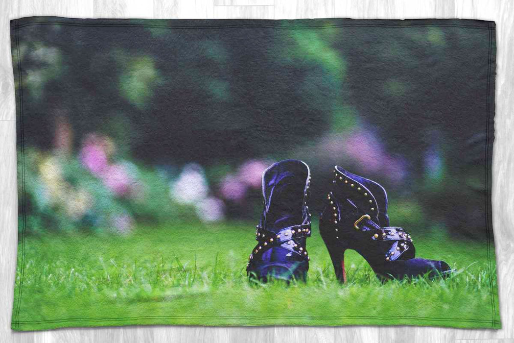 A blanket laid flat on a grey floor, the blanket having an image of a pair of purple high heel shoes sat on a grass lawn