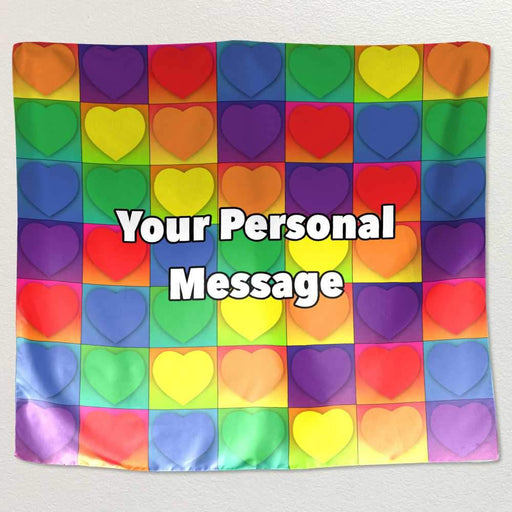 A tapestry hung on a white wall, the tapestry has lots of hearts on it, each heart inside a square, the hearts coloured with a rainbow colour, along with a personal message printed