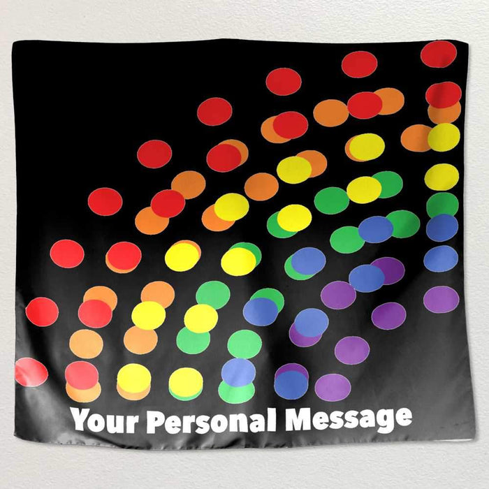 A tapestry hung on a white wall, the tapestry has a series of polka dots on it, the polka dots coloured with a rainbow colour on a black background, along with a personal message printed