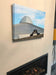 A canvas print of a pair of black high heel shoes sitting on a wall with a building in the background, the building has a slight UFO look to it, the canvas print is hung above a fire place in a living room