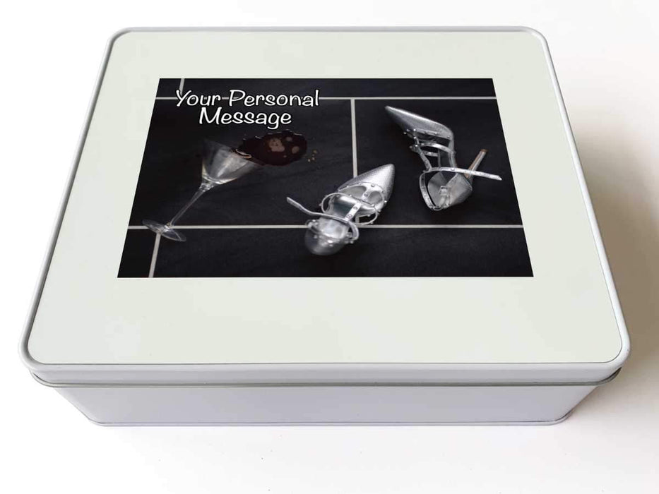 A tin box seen from top, the box having an image of a spilt cocktail next to a pair of silver high heel shoes, along with a personal message