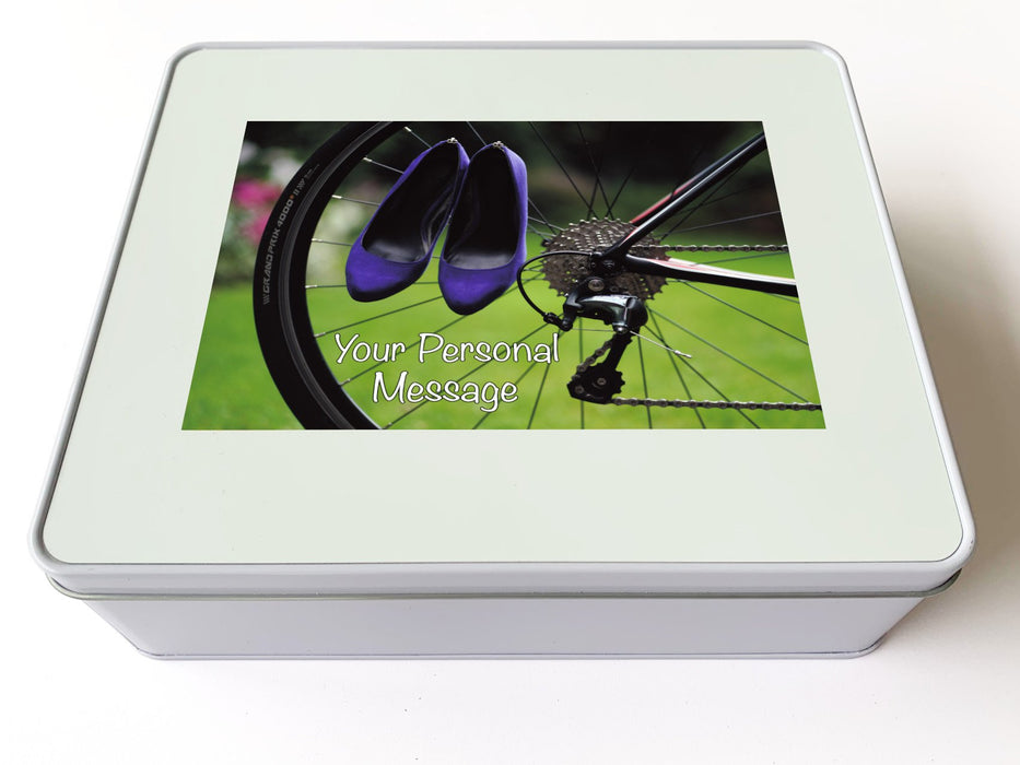 A white tin box, upon the lid is an image of a pair of purple high heel shoes hung on the rear wheel of a road bike, along with a personal message
