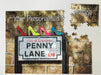 A jigsaw seen from the top, the jigsaw having an image of the famous penny lane road sign with four coloured shoes sat upon the road sign, along with a personal message, the jigsaw is broken into a few pieces