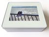 A white tin box with an image of a ocean with a pair of high heel shoes hanging on the railings in the foreground