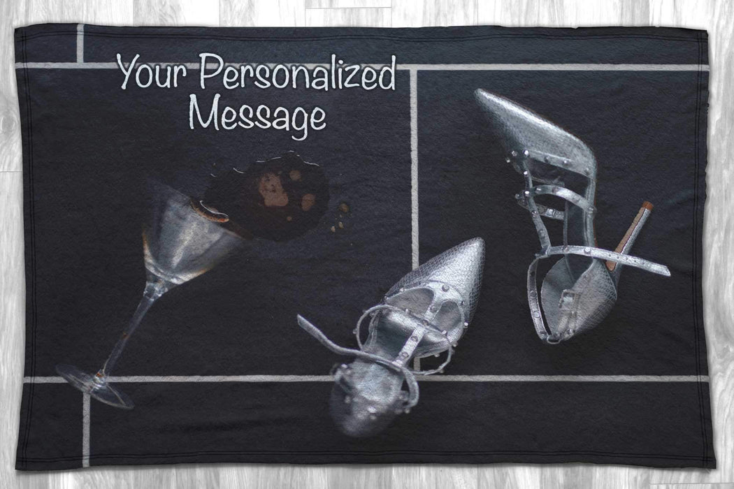 A blanket flat on the floor, the blanket has an image of a pair of silver shoes next to a spilt cocktail with a personal message printed