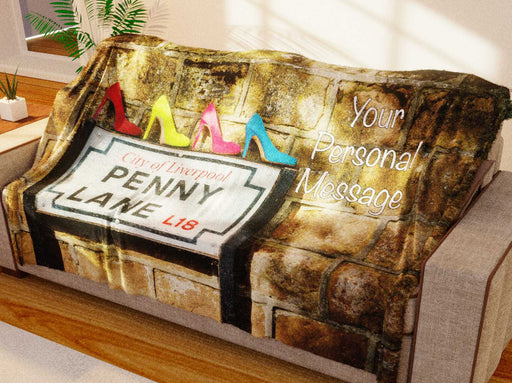 a fleece blanket draped over a couch in a living room, the blanket having an image of the penny lane street sign with four coloured shoes on the top, one in pink, one yellow, one blue, one red