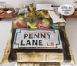 a duvet on a bed, the duvet having a picture of the penny lane road sign in liverpool with 4 coloured high heel shoes on top of the sign, part of duvet is folded back to reveal a white underside