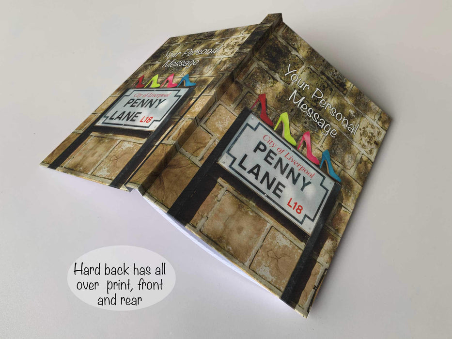 An open hardback notebook having a cover with an image the penny lane road sign in liverpool, with four high heeled shoe sitting on the sign, the shoes coloured red, blue, pink and yellow, and a personal message