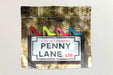 a tapestry fabric hung on a white background the tapestry showing the famous penny lane road sign with four coloured ladies high heel shoes upon it