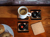 four coasters on a table each with image of a vinyl record playing on a record player, with a coffee cup on one coaster and some doughnuts in the back ground, with one coaster upside down revealing a cork underside