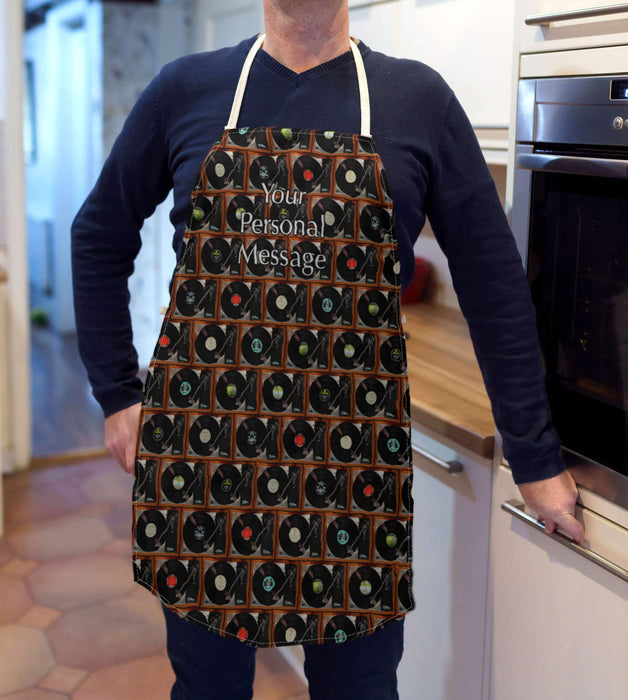 A man wearing an apron standing in a kitchen, the apron having a mosaic pattern of record players with a mixture of vinyl records and a personal message printed