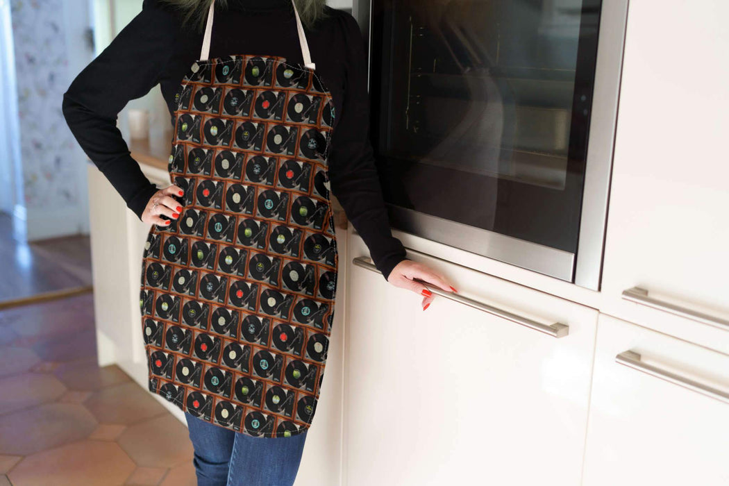 A woman wearing an apron standing in a kitchen, the apron having a mosaic pattern of record players with a mixture of vinyl records