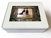 A white tin box, the lid having a photo on it, that photo is an image of a pair of orange and black high heel shoes reflected in a brass mirror, along with a printed personal message