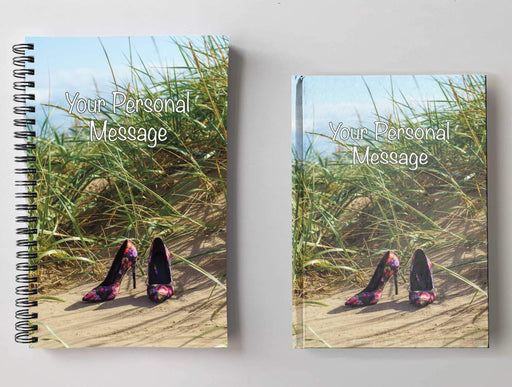 Two notebooks, side by side, both having covers with an image of a pair of multi coloured high heel shoes sat on a beach near a sandhill, with sandhill grasses in background, with a personal message on the cover