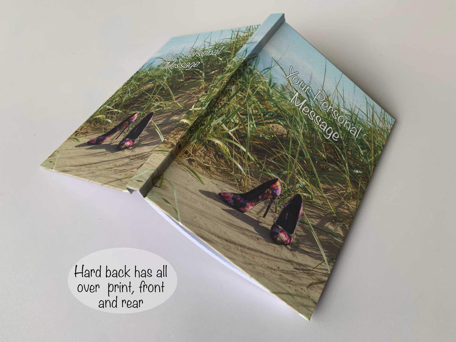 An open handback notebook with a cover of an image of a pair of multi coloured high heel shoes sat on a beach near a sandhill, with sandhill grasses in background, with a personal message on the cover