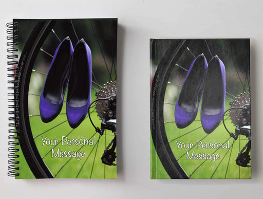 Two notebooks, side by side, both having an image of rear wheel of a racing bicycle with a pair of purple shoes hung on the spokes of the rear wheel, with a personal message on the cover