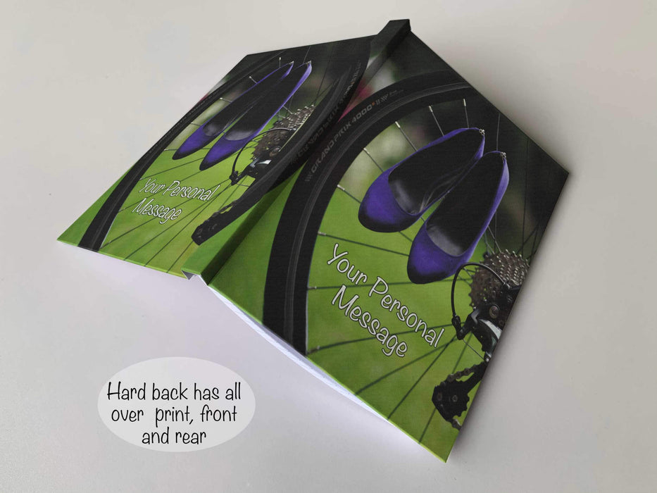 An open hardback notebook having a cover with an image of rear wheel of a racing bicycle with a pair of purple shoes hung on the spokes of the rear wheel, with a personal message on the cover