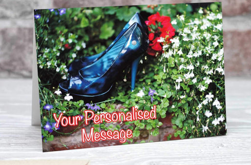 A birthday or greetins card showing a pair of blue high heeled ladies shoes placed in the middle of set of plants in a garden, the plants being mostly green leaves along with a large red rose and smaller white flowers