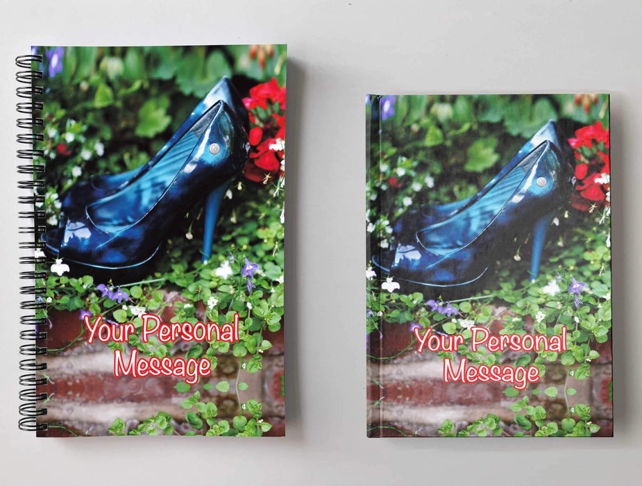 A pair of notebooks side by side, one spiral bound one hardback, both having an image on the cover of a pair of ladies high heel shoes resting inside a collection of outdoor plants along with a personal message
