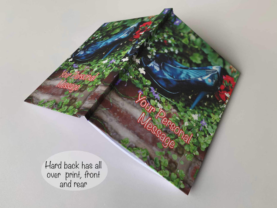 An open hardback notebook, the cover having an image on the cover of a pair of ladies high heel shoes resting inside a collection of outdoor plants along with a personal message