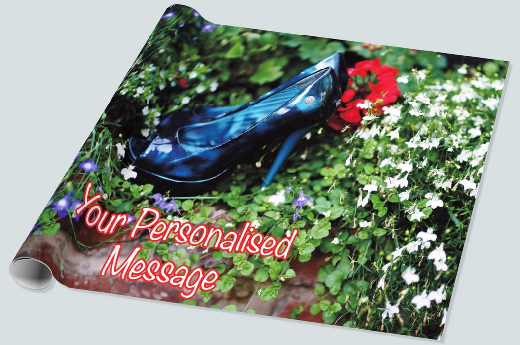 A roll of gift wrapping paper showing an image of pair of blue high heeled shoes n the middle of set of plants in a garden, the plants being mostly green leaves with a large red rose and smaller white flowers