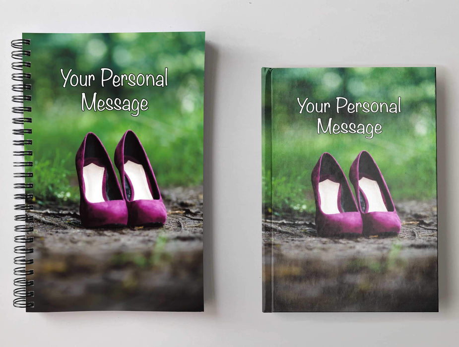 Two notebooks, side by side, both having covers with an image of a pair of purple high heeled shoes on a brown muddy footpath adjent to a grassy area along with a personal message on the cover