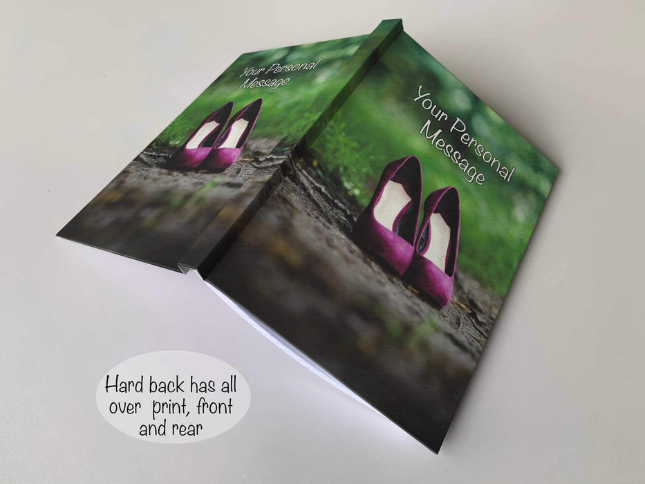 An open hardback notebook having a cover with an image of a pair of purple high heeled shoes on a brown muddy footpath adjent to a grassy area along with a personal message on the cover