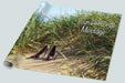 A roll of wrapping paper showing a pair of multicoloured high heel shoes on a beach in the sand hills, with sand hill grasses in close proximity to the shoes and a clear blue sky in the background