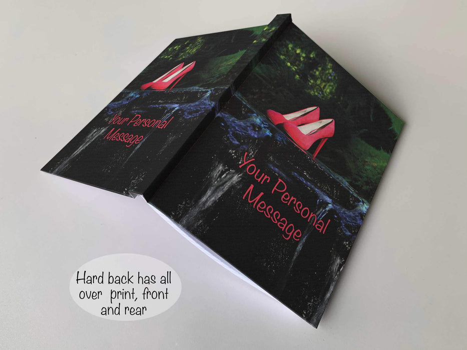 An open hardback notebook with a cover having an image of pink high heeled shoes on a rock midway across a flowing river, along with a personal message on the cover