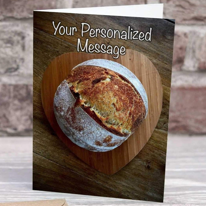 a birthday or greetings card on a desk with an envelope and pen, the card having an image of a sourdough loaf of bread sat on a wooden heart shaped tray