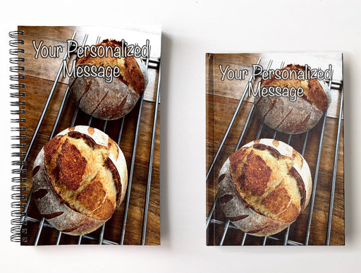 a pair of notebooks, one spiral bound, one hard back, next to each with image of two large sourdough loaves sat on a wire rack upon a table and seen from angled position