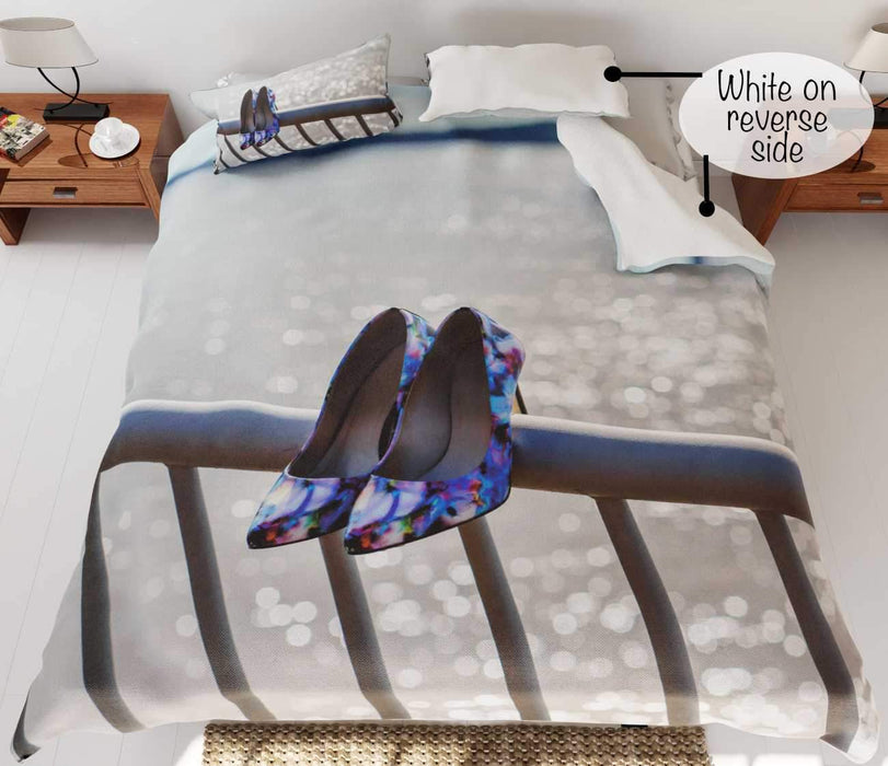 A duvet cover on a bed, the duvet having an image of a pair of purple high heel shoes hung on railings in front of the ocean, with part of the duvet folded to reveal a white underside
