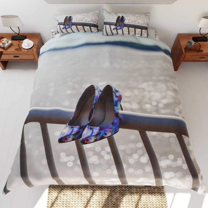 A duvet cover on a bed, the duvet having an image of a pair of purple high heel shoes hung on railings in front of the ocean
