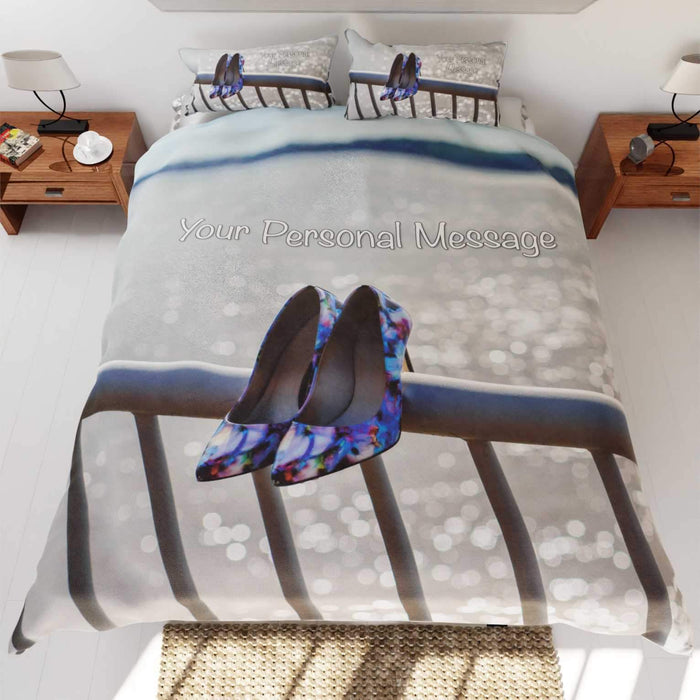 A duvet cover on a bed, the duvet having an image of a pair of purple high heel shoes hung on railings in front of the ocean along with a printed personal message