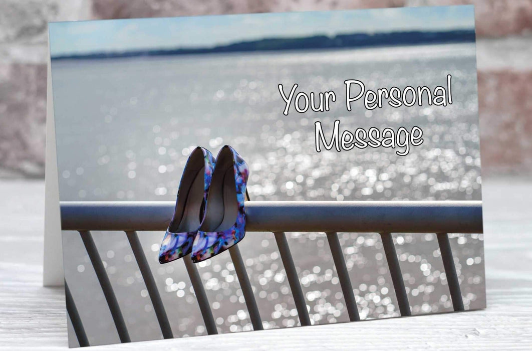 A birthday or greetings card showing an ocean with protective railings in the foreground, on the railings are a pair of blue and purple high heel shoes, there is a personal message on the card