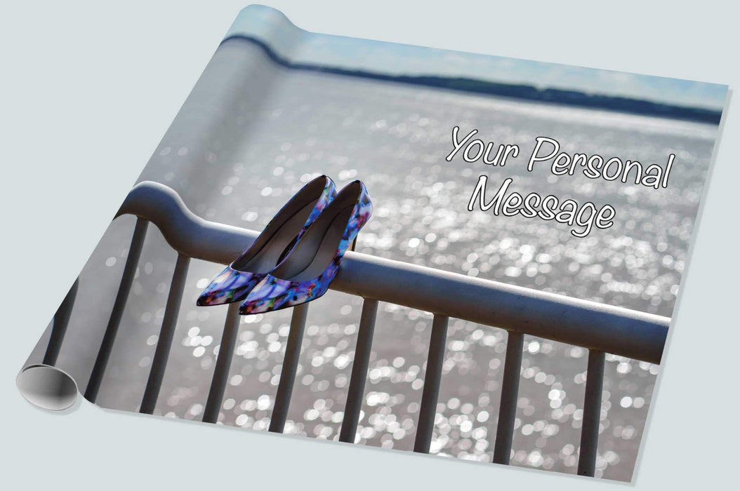 A roll of wrapping paper showing an ocean with protective railings in the foreground, on the railings are a pair of blue and purple high heel shoes, there is a personal message