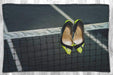 A blanket flat on a grey floor, the blanket having an image of a pair of yellow high heel shoes hung on the net of a tennis court